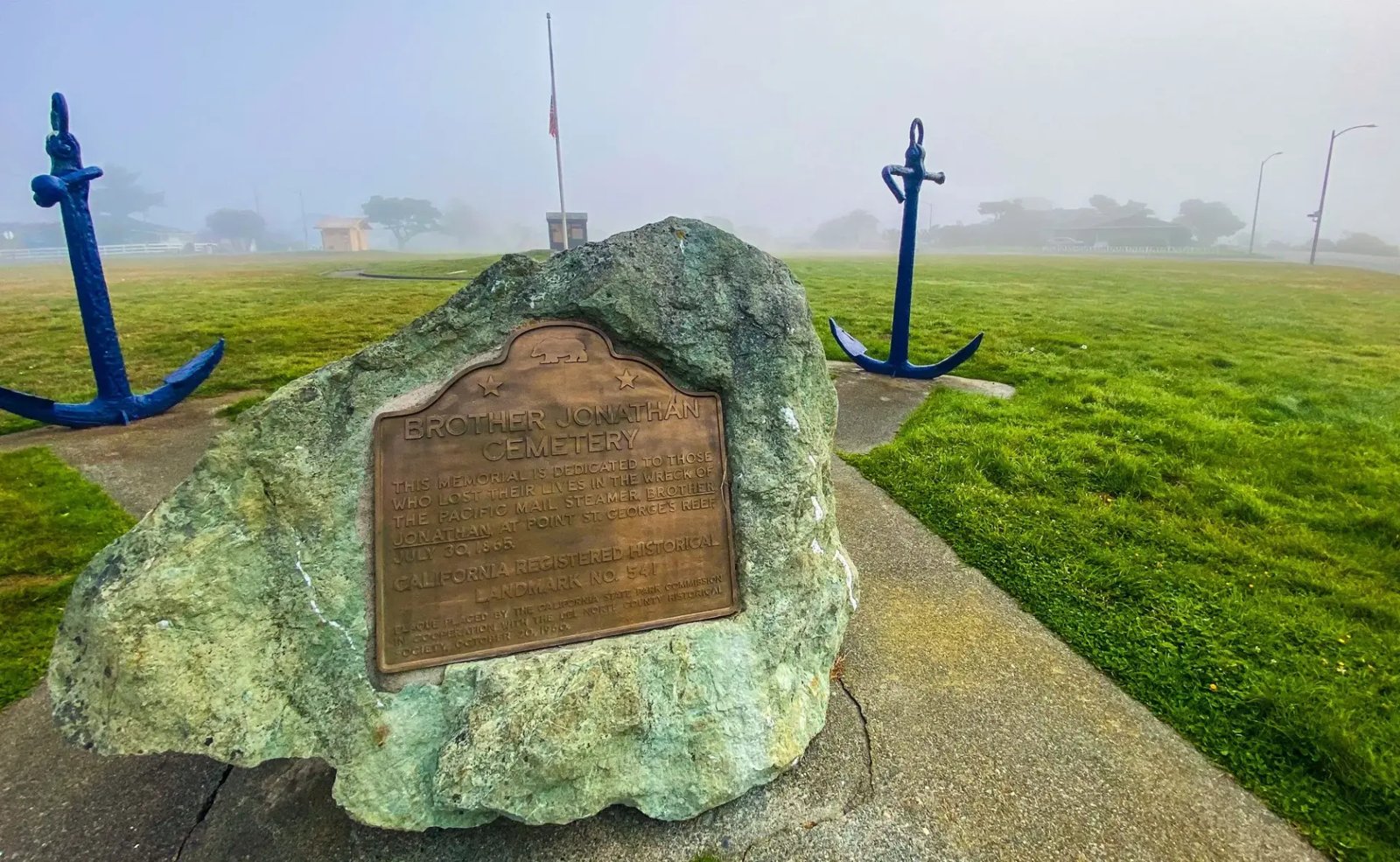 Bronze memorial plaque mounted on a boulder at the Brother Jonathan Cemetery in Crescent City, flanked by two blue anchors in coastal fog
